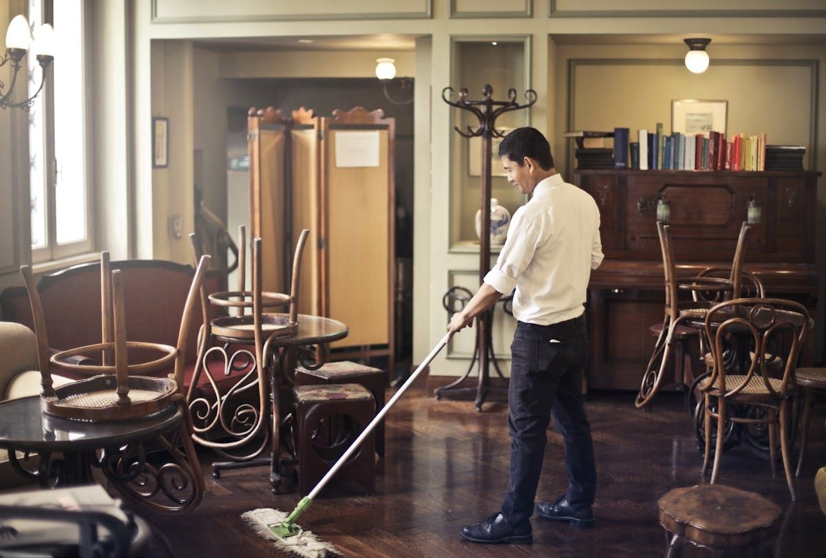 Man sweeping the floor in a cozy cafe with stacked chairs and tables.