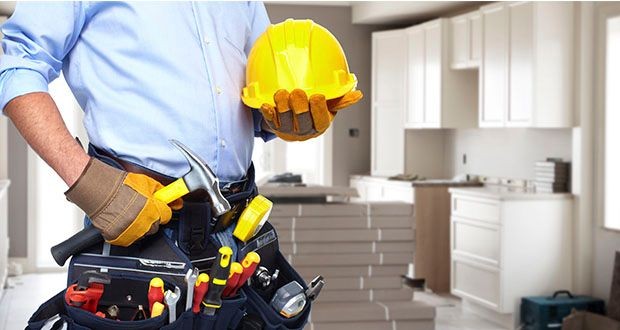 Construction worker holding a yellow hard hat and wearing a tool belt in a kitchen under renovation.