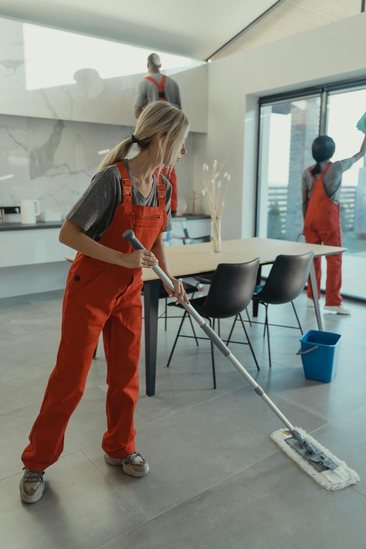 Three workers in red overalls cleaning a modern kitchen with mops and cloths.