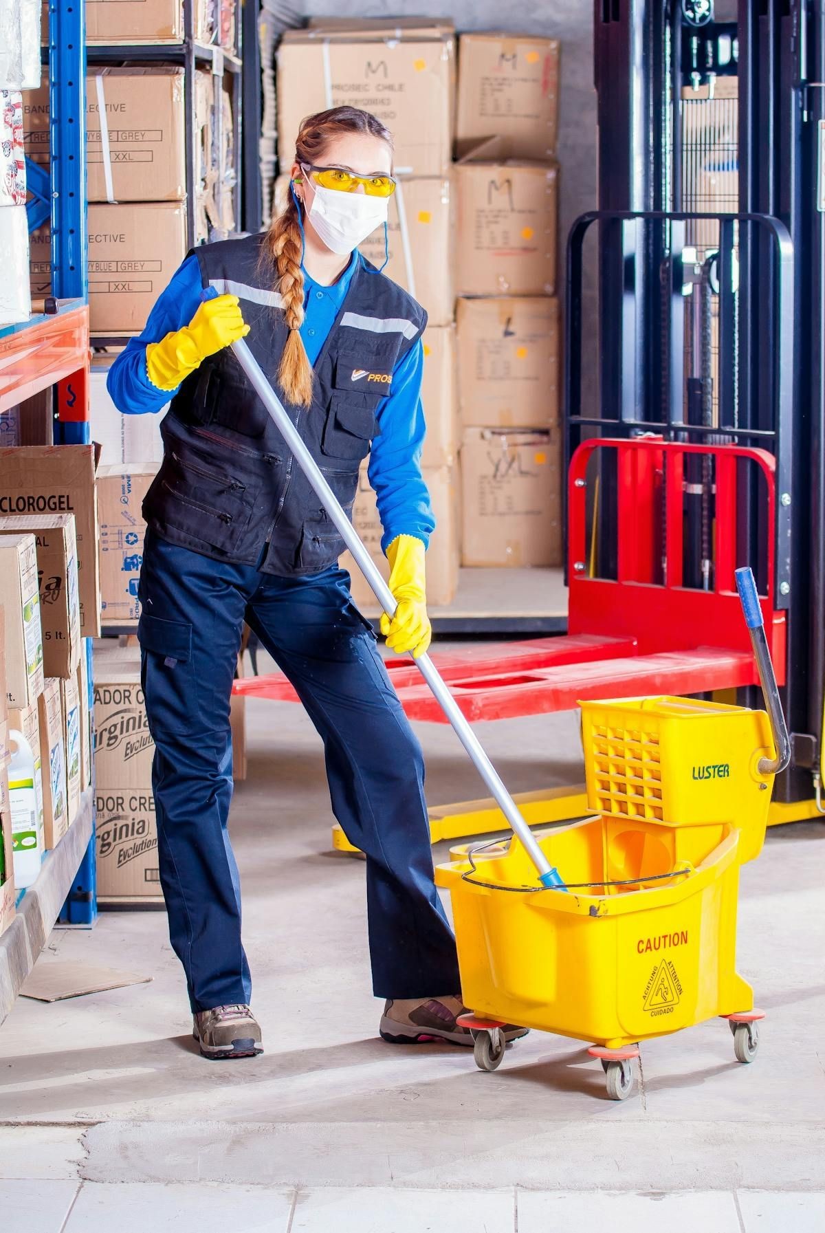 Person wearing protective gear mopping a warehouse floor with yellow caution mop bucket.