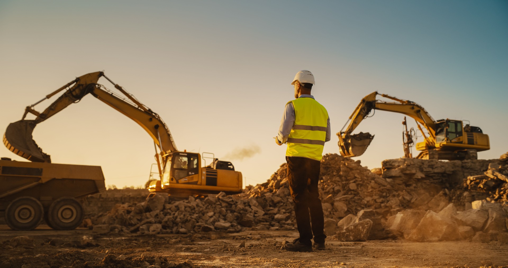 Caucasian Male Urban Planner Wearing Protective Goggles And Using Tablet On Construction Site On A Sunny Day. Man Inspecting Building Progress. Excavator Loading Materials Into Industrial Truck